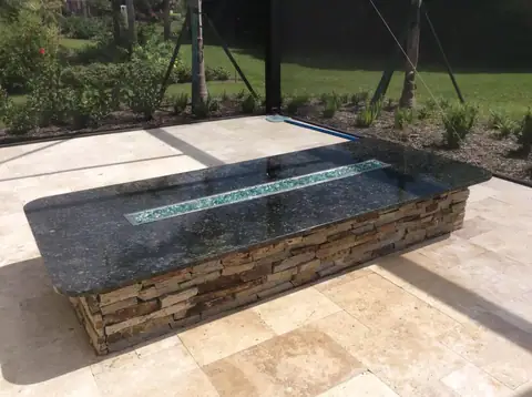 Custom outdoor linear firepit with black granite top and natural stone surround in Ft. Myers, FL. Custom outdoor linear firepit with black granite top and natural stone surround on travertine pool deck in the lanai.